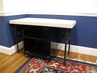 View of the Lifetime plastic folding table with black metal legs, positioned over the black rolling cart on a decorative rug with blue walls in the background.