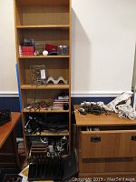 Tall wooden shelf with various items including ornaments, deck toy, thermometer, pens, briefcases, flashlights, and other small tools or electronics. To the right, a wooden surface holds multiple coiled electrical cords in white and brown.