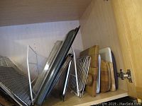 Photo of bakeware, cutting boards, wire racks and pans stacked upright in a wooden cabinet.