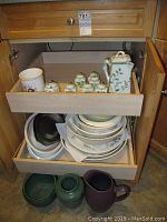 Wood cabinet open showing drawer with Andrea by Sadek ceramic set with tall pitcher, six small containers, and sugar bowl, and shelves below with stacked serving bowls and platters. Three ceramic planters on floor of cabinet: purple pitcher, green vase, green planter.