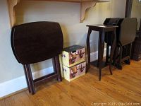 Set of four matching dark wood TV trays with stand, two small pedestal boxes, small square side table, and two additional TV trays leaning against wall.