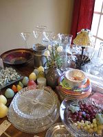 Photo showing assortment of glass bowls, vases, metal pitcher, faux flowers, candles, party plates, and lamp on a wooden table near window with red curtain.