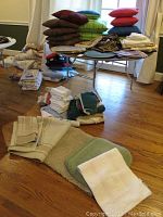 View of linens arranged on and around a folding table including stacked throw pillows in multiple colors, folded linens on the table, and assorted towels and rugs laid on the floor.