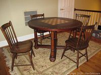 Round wooden game table with drop leaf sides and chess/checkerboard pattern on top, four matching wooden Hitchcock chairs around it, placed on a floral patterned area rug on a hardwood floor.