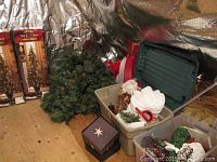 Various Christmas decorations spread on and around a plywood floor against a reflective foil wall, including greenery garland, Santa figurine, small decorative trees, a plastic storage bin with additional decor items, a miniature wrapped-sleigh with packages, and a basket.