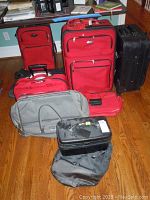 Image showing the full assortment of luggage items on a hardwood floor near furniture. Includes multiple red soft-sided suitcases, a grey duffle bag, black suitcase and smaller black cases.