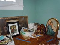 Wide shot showing assortment of religious items on wooden table, including framed prints, crosses, rosaries, and small statues.