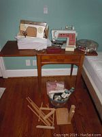 Full view of two Singer sewing machines placed on a wooden table with a brown finish, including the storage drawer beneath. Additional sewing accessories shown nearby on the floor beside the table.