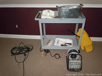 Metal utility cart holding paint supplies, buckets, utility light positioned on floor, and extension cords on floor around cart.