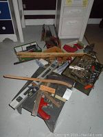 Overall assortment of tools including levels, rulers, saw, wrenches, vice, broom, and drill bits displayed on the floor