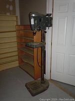 Full view of the Sears Craftsman drill press standing upright beside a wooden shelf and nearby door in a workshop or storage area.