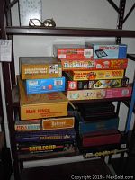 Photo of board games and puzzles stacked on metal shelving showing titles including Battleship, Mahjong, Pass Word, Yahtzee, Parcheesi, The Saturday Evening Post puzzle, and Snow & Go game.
