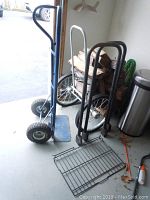 Photo showing blue Angelus dolly, black metal two-wheeled cart, and metal grate parts for firewood rack on garage floor near firewood stack and trash can.