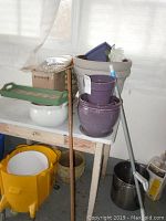 View of assorted ceramic and plastic garden pots of various sizes and colors arranged on a table and floor, including large beige pot, purple pots, yellow plastic pot, and some garden tools leaning nearby.