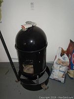 Full view of black Weber smoker showing dome lid, cylindrical body, door opening, and lower firebox compartment, alongside bags of charcoal or wood chips.
