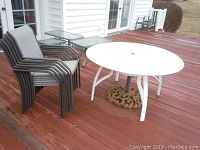 Full view of round glass top patio table with umbrella holder base and six stacked metal mesh chairs on wooden deck