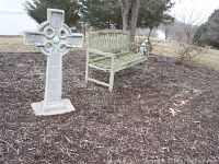 Full view of cement Celtic cross and adjacent wooden bench in a garden area to show scale and outdoor setting.
