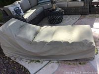 Outdoor wicker chaise lounge covered with beige protective cover, placed on stone patio with surrounding furniture.