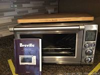 Front view of stainless steel toaster oven with instruction manual propped in front and bamboo cutting board atop oven.