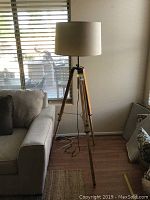 Full view of the wooden tripod floor lamp with beige drum shade standing beside a gray sofa in a room with hardwood floor and window blinds.