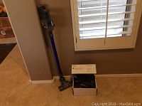 Dyson DC44 Animal cordless stick vacuum standing upright with blue tube and floor cleaning head, box of spare attachments on the floor beside it.