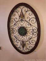Front view of oval metal wall art with brown hammered frame, black scrollwork, gold leaf accents, and green flower center.