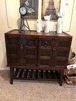 Front view of dark wooden cabinet showing four doors with carved decorative panels and metal ring pull handles. Decorative items placed on top.