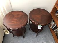 Two round brown wooden side tables with smooth polished tops and lower shelves, positioned side by side on carpeted floor near window and bookshelf.