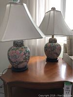 Two Asian-inspired table lamps with floral ceramic bases on a wooden table by a window.