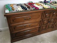 Frontal view of wooden dresser showing three drawers on left and wooden paneled double doors on right, glass top visible with assortment of jewelry laid out.