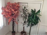 Two artificial trees side by side: autumn tree with orange-red leaves on left, green palm tree on right, white flowered tree in back not for sale.