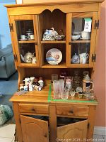 Full view of wooden kitchen hutch showing two glass doors, two open shelves, two drawers, and two cupboard doors below. Hutch filled with various glassware and ceramics included in photos but not part of the lot.