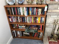 Wooden bookshelf with three shelves filled with multiple assorted hardcover and softcover books in good condition, visible book titles of diverse genres.