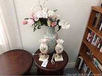 Full view of floral vase with large artificial flowers and two matching small table lamps on round wooden tables.