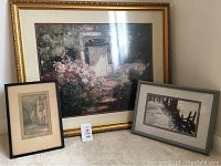 Three framed art pieces displayed on carpet: large floral/garden artwork with gold frame, smaller sepia street scene in black frame, smaller black and white waterfront boats scene in gray frame.