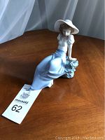 Porcelain figurine of a seated girl wearing a blue dress and white wide-brimmed hat, sitting beside a rock with a bird. Figurine is approximately 6 inches tall.