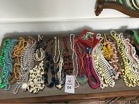 Overhead view of various colorful beaded necklaces laid out on a wooden surface including turquoise, gold, white, black and multicolor strands.