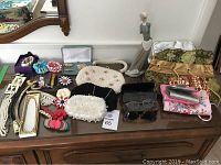 Wide view of lot items arranged on wood dresser including jewelry, clutches, fabric pouches, eyeglasses, and porcelain figurine.