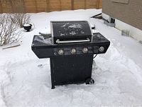 Front view of black Grillmaster propane BBQ grill with silver lid and four control knobs, placed outdoors on snow.