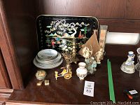 Asian inspired decorative lot on wooden shelf including black lacquer serving tray, metal plates, bowls, several figurines, a fan in box, metal compact with mirror, and various small decorative items