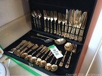 Open black storage chest containing the gold-tone cutlery set with forks, knives, spoons secured by elastic bands