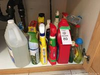 View of assorted cleaning items on cabinet shelf including spray bottles, gallon container, mousetraps, SOS pads, dish soap, scouring pads.