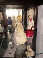 Two Royal Worcester lady figurines on a display shelf, one in a ruffled pastel dress and bonnet, the other in a white coat with red skirt and hat.