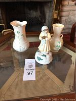 Wide photo showing all three items on glass table in front of fireplace: Schmid musical girl figurine in center, flanked by two Beleek vases with shamrock decoration.