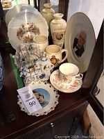 Photo showing several collector plates, mugs, teacup and saucer, a vase, and a blue and white ceramic figurine set on a wooden side table.
