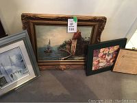 Four framed artworks displayed leaning against a wall: large coastal oil painting in ornate gold frame, floral print in green frame, vintage text print, and a winter scene print in silver frame.