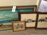 Four framed artworks leaning against wall on floor: large seascape painting at left, two black ink sketches of rural houses, and one small framed industrial scene painting in gold frame.