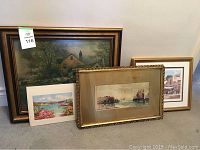 Four assorted framed artworks arranged on the floor showing diverse scenes including a house landscape and market scenes.