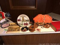 Photo of various decorative plates, including a Goldboro plate, carved plaques, a painted circular plate, a miniature orange wagon lamp, rollerblades, and a red hardcover book.