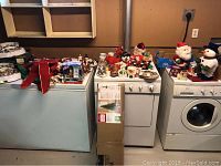 Wide shot showing assortment of Christmas decorations and collector plates arranged on top of washer and dryer units with prelit artificial tree box in front.